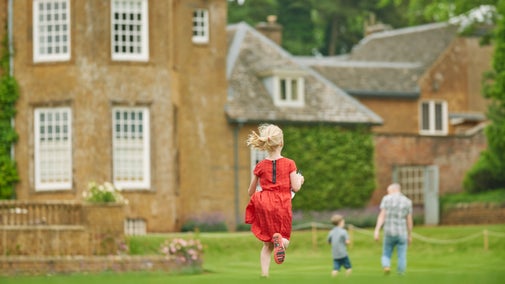 A girl in a red dress runs after a man and a small child across the lawn at Upton House, Warwickshire